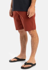 Rust-colored shorts with a straight cut, made of cotton, paired with black flip-flops. Features flat seams and no visible fasteners.