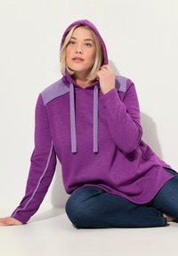 Sudadera larga morada con cordones elásticos, acentos morados en los hombros y aberturas laterales. Llevada con vaqueros oscuros, sentada sobre un fondo claro.