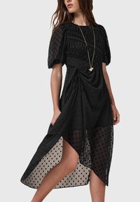 Vestido negro con mangas abullonadas, cuerpo fruncido y dobladillo asimétrico. Hecho de una tela texturizada con una capa transparente y un sutil patrón geométrico.