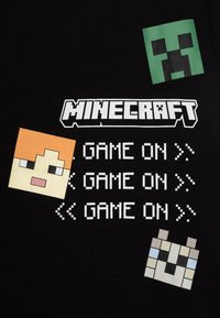 Melna auduma t-krekls ar četriem pikselētiem sejiem no Minecraft, ar tekstu "MINECRAFT GAME ON" baltā, treknrakstā.