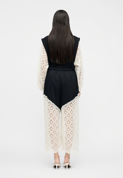 Femme aux longs cheveux noirs portant un haut noir sans manches par-dessus un chemisier en dentelle blanche à manches longues et un pantalon en dentelle assorti, des mules blanches à talons.