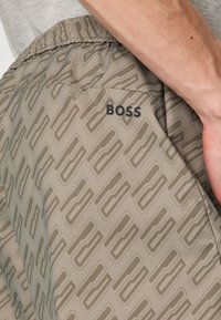 Shorts gris avec un motif géométrique en relief répétitif dans une teinte plus foncée. Le nom de la marque "BOSS" est imprimé en noir sur le côté.