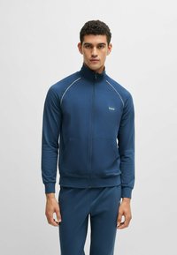 Giacca a track con zip color blu navy realizzata in tessuto morbido, con colletto alto, dettagli in piping bianco e un logo sul lato sinistro del petto.