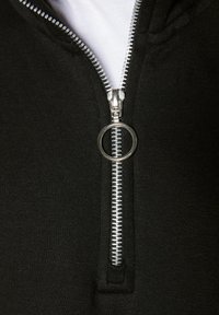 Detalle de sudadera con cremallera negra que presenta una cremallera plateada con un tirador circular, fabricada en tejido suave que muestra una textura lisa.