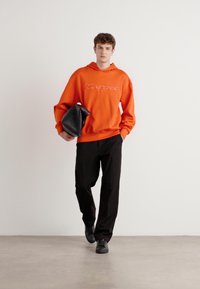 Felpa arancione con logo, pantaloni neri e borsa nera. Il modello si trova davanti a uno sfondo chiaro, mostrando un abbigliamento e accessori casual.