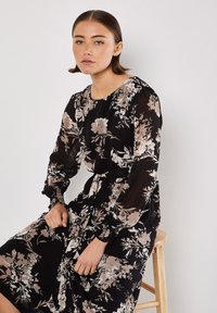 Schwarzes Blumenkleid mit langen, transparenten Ärmeln, das ein Muster aus hellrosa Blumen und eine taillierte Taille für eine definierte Form und Textur aufweist.