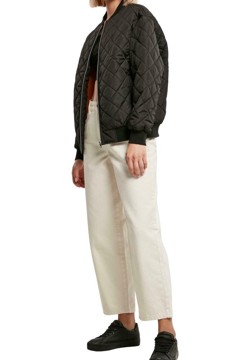 Femme portant une veste bomber matelassée noire, un haut noir, un pantalon large blanc taille haute et des baskets noires, debout devant un fond blanc.