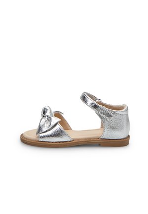 Sandali metallici argentati con dettaglio a fiocco, punta aperta, cinturino alla caviglia e suola piatta con base marrone chiaro.