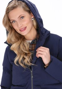 Femeie cu păr blond, purtând cercei pătrați argintii și o geacă puf de culoare albastru închis cu glugă, ținând geaca aproape de fermoar.