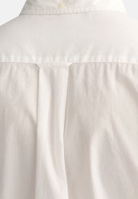 Bílá bavlněná košile s límečkem na knoflíky. Obsahuje středový sklady vzadu a hladkou, měkkou texturu s jemným leskem.