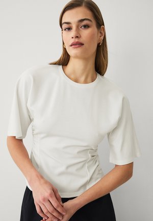 Kvinde med glat brunt hår iført løs hvid kortærmet top, guldringe øreringe og sorte bukser, poserer mod en ensfarvet baggrund.