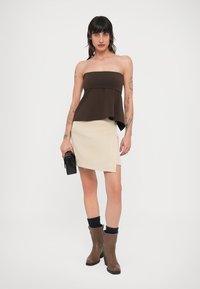 Καφέ τοπ χωρίς τιράντες με φουσκωτό τελείωμα, συνδυασμένο με ασύμμετρη μίνι φούστα σε κρεμ απόχρωση. Το μοντέλο φοράει καφέ μποτάκια μέχρι τον αστράγαλο και κρατάει ένα μαύρο τσαντάκι χειρός.