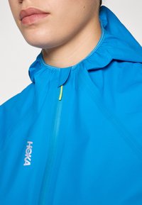 Blue lightweight jacket with a cinched collar, half-zip closure, and a small logo on the left side. Smooth texture, water-resistant fabric.
