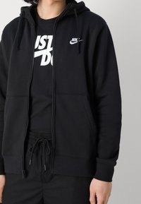 Svart zip-up hoodie i mjukt material, med en framficka och huva med dragsko, samt en liten Nike-logga på bröstet.