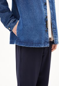 Denim jack in diepblauw, met drukknoopsluitingen, een puntige kraag en zijzakken. Gedragen over donkere, gladde stoffen broeken.