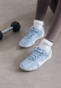 Könnyűkék sportcipőt és fehér zoknit viselő személy ül egy szürke padlón egy fekete súlyzó mellett.