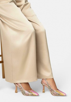 Bein in beigen Hosen mit weitem Bein, sitzend auf einem Holzstuhl, getragen werden mehrfarbige Slingback-Pumps mit Schlangenledermuster in Pink, Gelb und Braun.