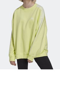 Žlutá oversized mikina s bílým logem Adidas a třemi bílými pruhy na rukávech. Měkká tkanina, žebrované manžety a lem. Uvolněný střih.