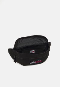 Bolsa de cintura de tela negra con cremallera superior, que presenta un parche de logo en rojo, blanco y azul, y un pequeño bolsillo interior. El interior está forrado en negro.