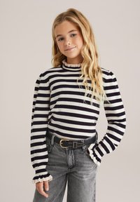 Tricou cu mânecă lungă în dungi, de culoare bleumarin și crem, cu guler înalt cu volane și mâneci bufante; asortat cu blugi gri cu talie înaltă.