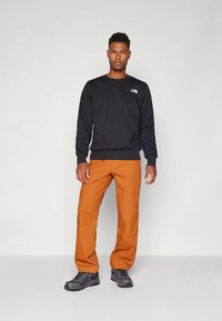 Muž stojaci, oblečený v čiernej dlhorukávovej košeli, oranžových nohaviciach a sivých turistických topánkach na jednotnom bielom pozadí.