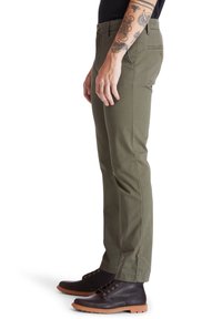 Man wearing olive green slim-fit pants and dark brown leather lace-up boots, showing tattooed arm and side profile.