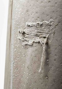 Gros plan sur un tissu beige avec des gouttes d'eau et une déchirure horizontale effilochée montrant des fils usés.