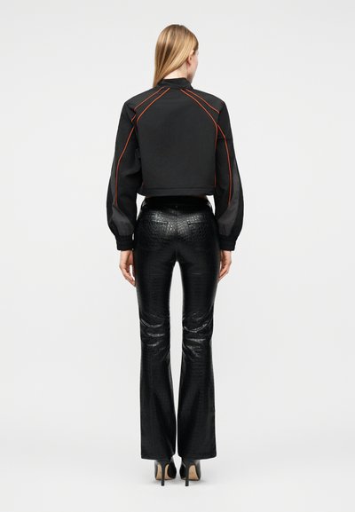 Γυναίκα με ξανθά μαλλιά που φοράει ένα κοντό μαύρο μπουφάν με πορτοκαλί ραφές και μαύρο υφασμάτινο παντελόνι με ψηλή μέση και τακούνια, γυρισμένη από την άλλη πλευρά.