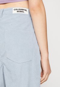 Nära bild av en person som bär ljusblå kordflorsbyxor med en etikett "COLOURFUL REBEL" på midjan och en lavendelfärgad topp.