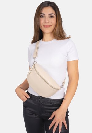 Beige Kunstleder Umhängetasche mit einer geschwungenen Form, Reißverschlussfach und verstellbarem Tragegurt, getragen über einem weißen Hemd.