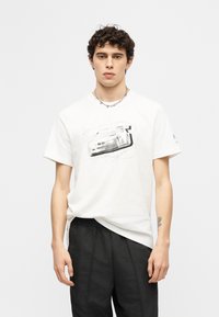 Νέος άνδρας με σγουρά μαλλιά φοράει λευκό T-shirt με γκριζο-scale γραφικό αυτοκινήτου, μαύρο παντελόνι και ασημένιο αλυσίδα κολιέ σε απλό φόντο.