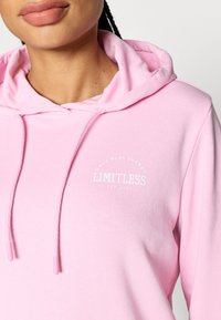 Light pink hoodie featuring a kangaroo pocket, drawstrings, and a white printed logo with rounded text near the neckline. Soft fabric texture.
