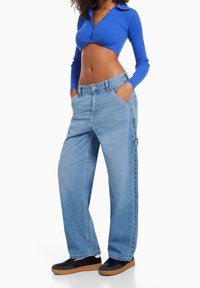 Kobieta w krótkim, niebieskim topie z długim rękawem, w jasnoniebieskich, luźnych spodniach z wysokim stanem oraz czarnych sneakersach z gumową podeszwą, z rękami w kieszeniach.