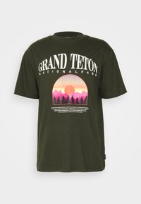Camiseta de algodón verde oliva con un gráfico redondo que muestra montañas rosas y moradas, un sol y texto blanco que dice "Parque Nacional Grand Teton."