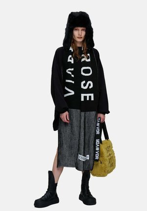 Femme portant un chapeau trappeur noir en fourrure, un pull noir oversize, une jupe rayée, des bottes noires, une grande écharpe noire et blanche, et tenant un sac en fourrure jaune.