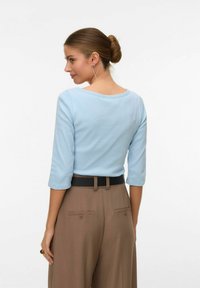 Vrouw met haar in een knot, gekleed in een lichtblauw topje met driekwart mouwen, ingestopt in een hooggetailleerde bruine broek met een zwarte riem.