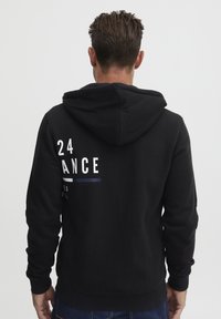 Melna kapuce ar kengura kabatu, strukturēta auduma, un ar drukātu dizainu, kas aizmugurē redzams ar "24" un "DANCE" baltā un zilā krāsā.