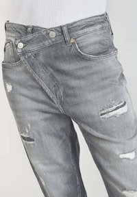 Harmaat farkkushortsit kuluneella viimeistelyllä, joissa epäsymmetrinen vyötärösuunnittelu, näkyvät saumat ja useita taskuja.