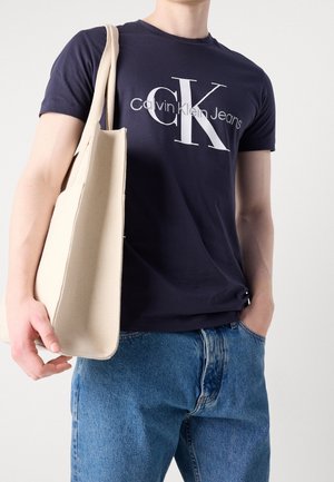 Mand iført navy Calvin Klein Jeans t-shirt og blå jeans, der holder en beige tote-taske på skulderen.