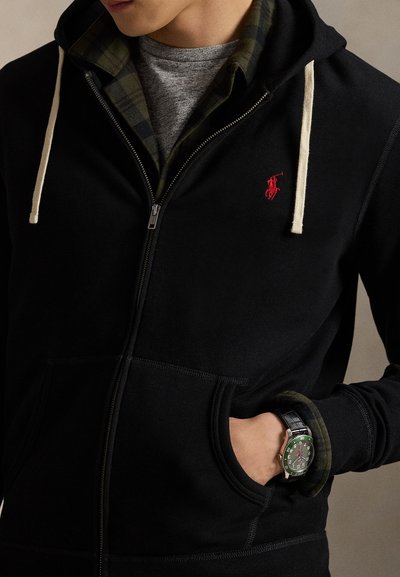 Moški, ki nosi črno kapucar s patentnim zadrgo in belimi vrvicami ter karirasto srajco pod njim, roko v žepu, ki kaže zeleno zapestno uro.