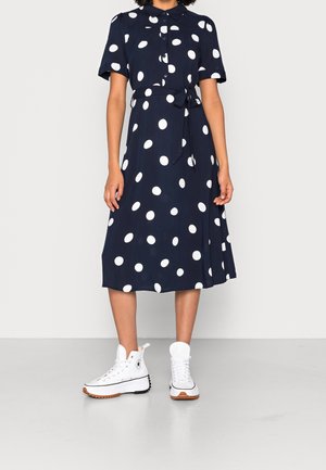 Femme portant une robe midi boutonnée bleu marine à pois blancs, manches courtes, ceinture à la taille, et baskets montantes blanches.