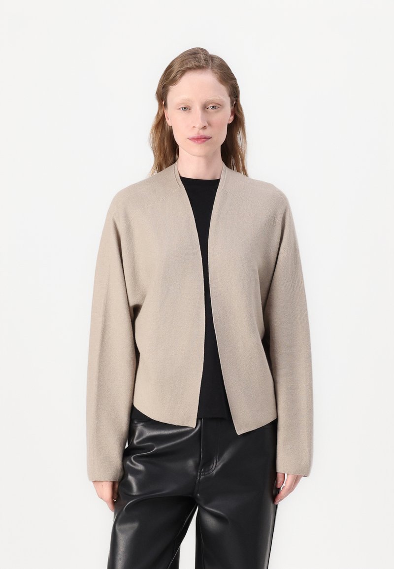 Beige cardigan med en avslappet passform, åpen front og lange ermer, laget av myk teksturert stoff. Båret over en svart topp og svarte bukser.