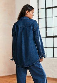 Niebieska dżinsowa koszula overshirt, z kołnierzykiem, długimi rękawami i okrągłym hemem. Materiał ma teksturowane wykończenie z widocznymi detalami ściegów.
