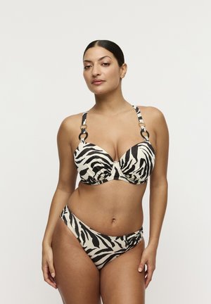 Vrouw staand in een zwart-witte bikini met zebraprint en decoratieve ringen aan de bandjes, voor een effen witte achtergrond.