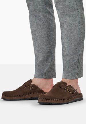 Zoccoli slip-on in suede marrone con design cucito, fibbia regolabile e suola in gomma testurizzata, abbinati a pantaloni grigi a gamba affusolata.