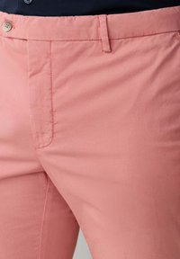 Primo piano di una persona che indossa pantaloni rosa corallo con bottone e passanti per cintura, abbinati a una maglia scura.
