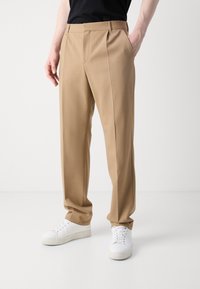 Beige pielēti bikses ar gludu tekstūru, kas aprīkotas ar divām priekšējām krokām, sānu kabatām un brīvāka piegriezuma, saskaņotas ar baltām sporta apaviem.