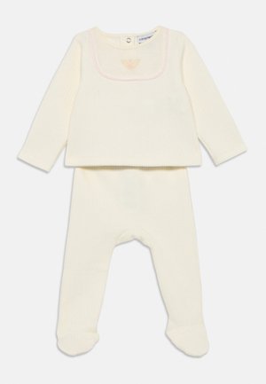 Elfenbeinfarbener, gerippter Baby-Body mit langen Ärmeln, Fußteil, rundem Ausschnitt und kleinem, rosa gesticktem Adler-Logo auf der Brust.