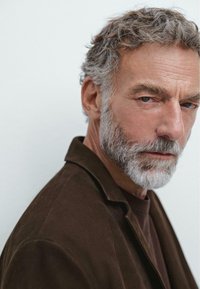 Ritratto di un uomo con capelli grigi corti e mossi e una barba, che indossa una giacca di pelle scura sopra una camicia scura, su uno sfondo chiaro.