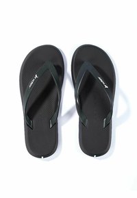 Chanclas de goma negras con una base texturizada. Las correas verdes presentan un patrón diagonal. Logo blanco impreso en el lateral.